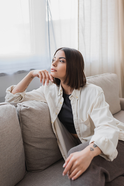 Young tattooed woman looking away while sitting on couch at home