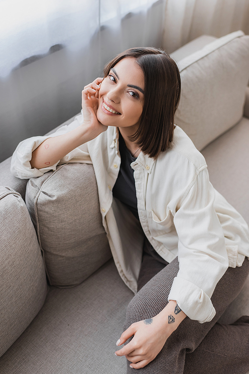 Overhead view of smiling tattooed woman looking at camera while sitting on couch at home