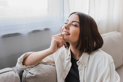 Cheerful young woman in shirt looking away while sitting on couch at home