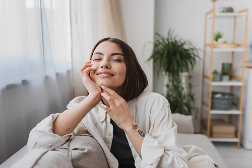 Overjoyed tattooed woman touching face while sitting on couch at home