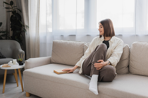 Smiling young woman taking book while sitting on couch at home