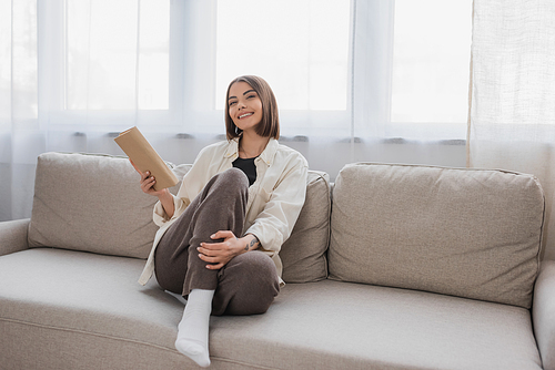 Cheerful young woman holding book while sitting on couch in living room