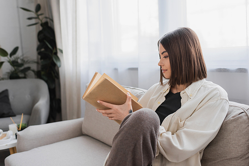 Side view of brunette woman holding book while sitting on couch at home