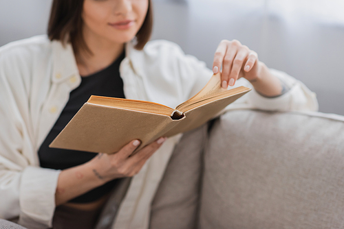 Cropped view of blurred woman reading book while sitting on couch at home