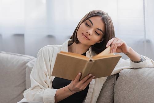 Pleased tattooed woman reading blurred book in living room