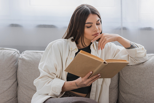 Tattooed young woman reading book on couch in living room