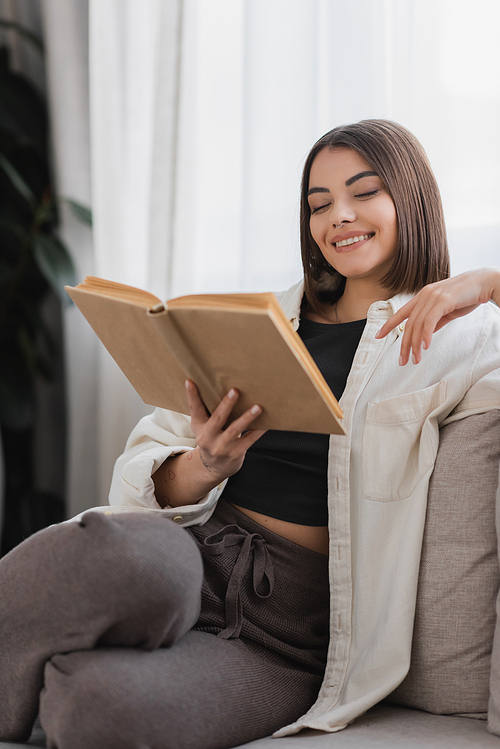 Positive woman reading book while resting on couch at home