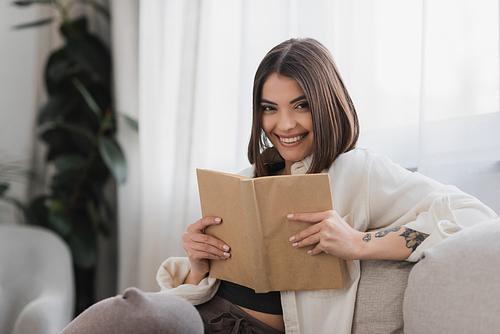 Positive tattooed woman holding book and looking at camera in living room