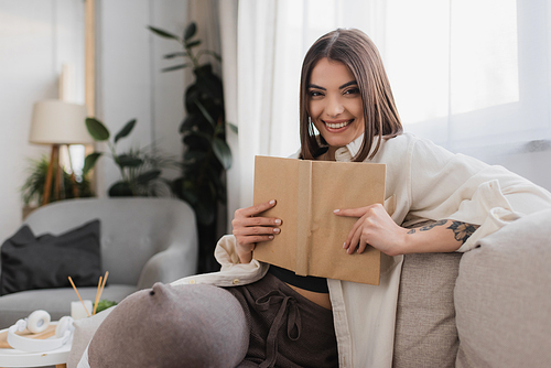 Smiling brunette woman holding book and looking at camera on couch at home