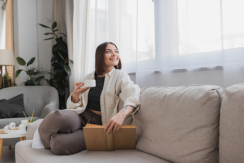 Positive woman holding cup of coffee and book while resting on couch at home