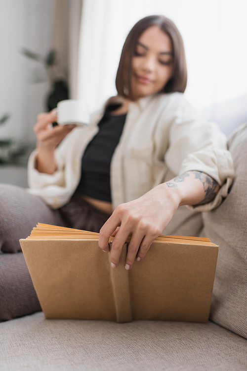 Blurred woman holding cup while reading book in living room