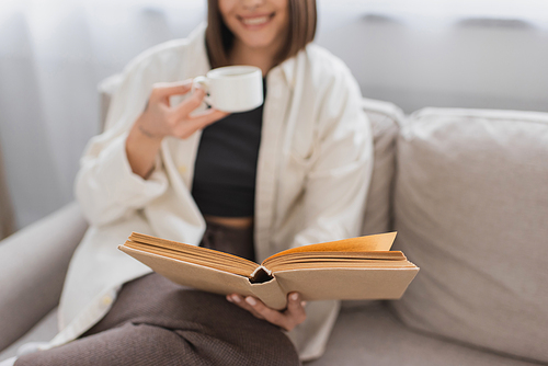 Cropped view of blurred woman holding coffee cup and reading book on couch at home