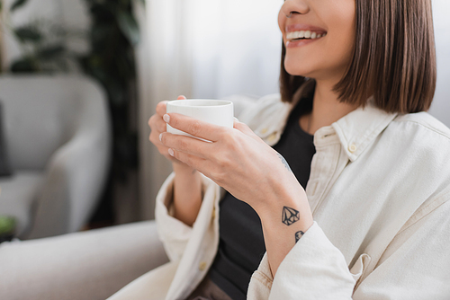 Cropped view of carefree tattooed woman holding cup of coffee in living room