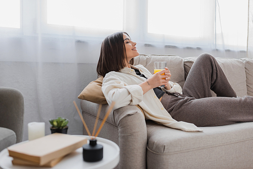 Side view of pleased woman holding glass of orange juice on couch at home