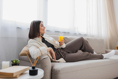 Side view of smiling brunette woman holding glass of orange juice while sitting on couch at home