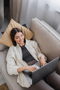 Top view of cheerful brunette woman using laptop while lying on couch at home