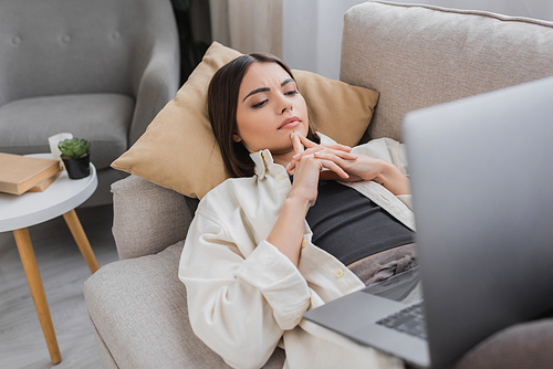 Focused brunette freelancer looking at laptop while lying on couch at home