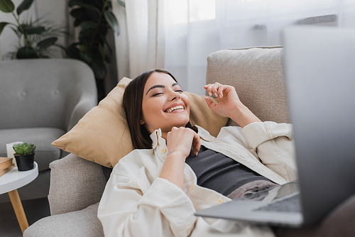 Cheerful brunette woman with blurred laptop while lying on couch at home