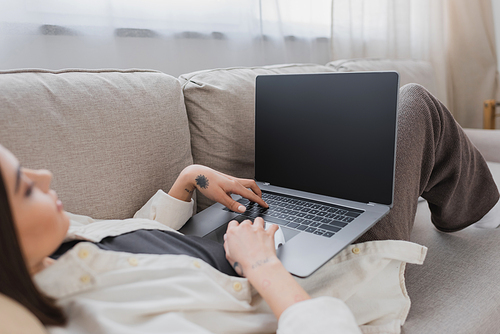 Blurred tattooed freelancer using laptop with blank screen on couch at home