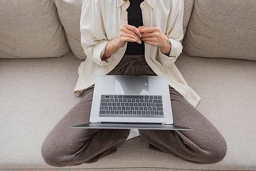 Cropped view of tattooed woman in casual clothes holding laptop while sitting on couch at home