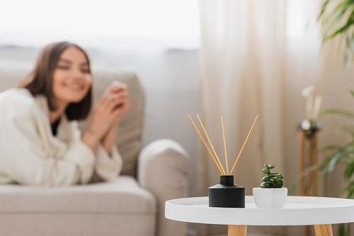 Plant and bamboo scented sticks on coffee table near blurred woman on couch at home