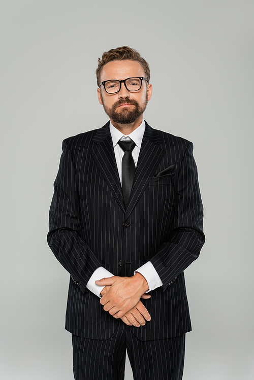 confident and bearded businessman in formal wear and glasses looking at camera isolated on grey