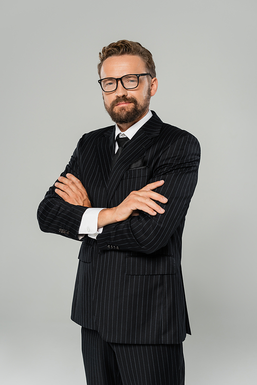 cheerful businessman in formal wear and glasses posing with crossed arms isolated on grey