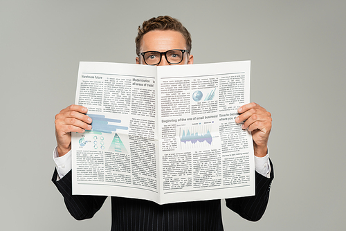 businessman in glasses looking at camera while covering face with newspaper isolated on grey