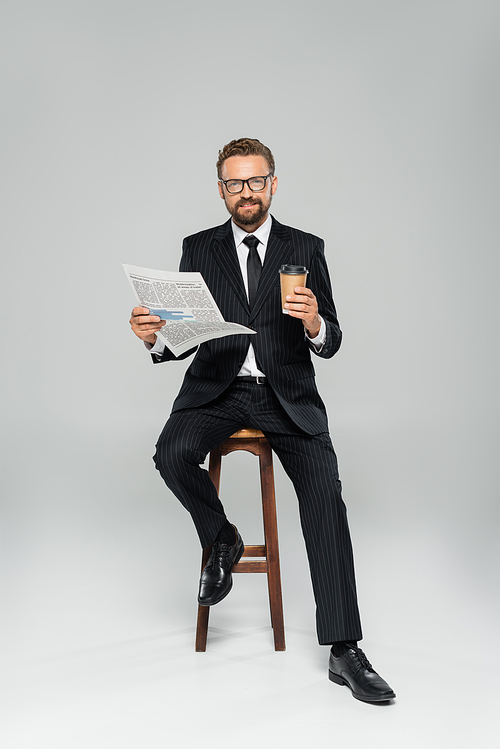 full length of happy businessman in suit and glasses holding newspaper while sitting with paper cup on grey