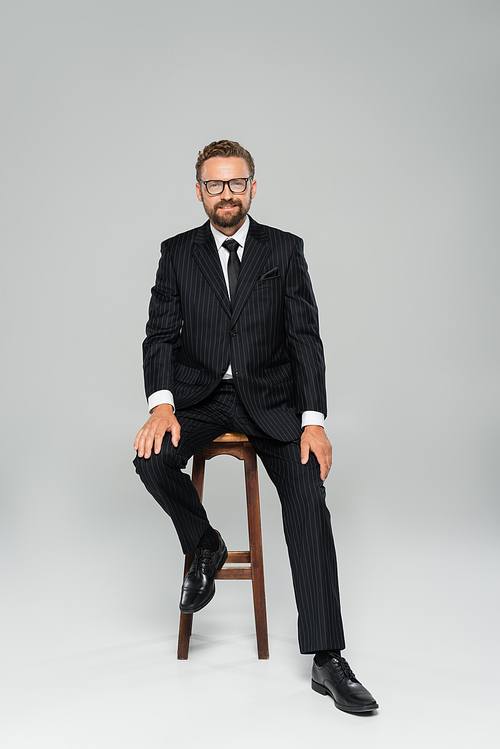 full length of happy businessman in suit and glasses sitting on wooden high chair on grey