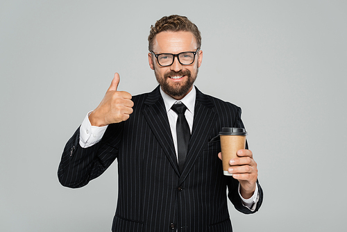 happy businessman in glasses and suit holding paper cup and showing thumb up isolated on grey