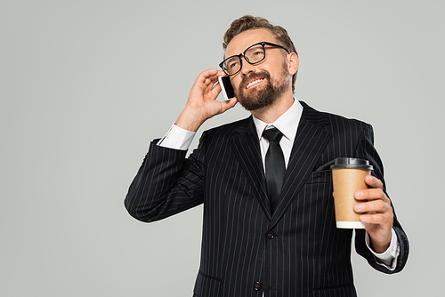 happy businessman in glasses and suit talking on smartphone and holding paper cup isolated on grey