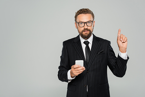 bearded businessman in glasses and suit holding smartphone and pointing with finger isolated on grey