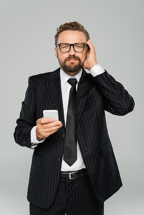 bearded businessman in glasses and suit having headache while holding smartphone isolated on grey