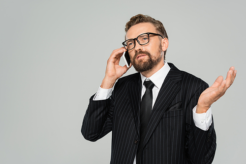 bearded businessman in suit and glasses talking on smartphone isolated on grey