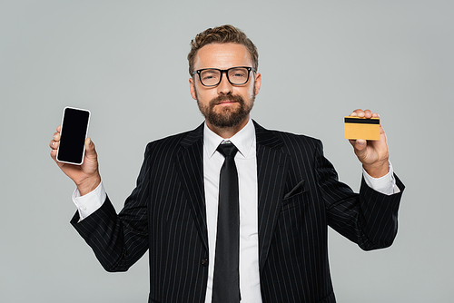 bearded businessman in suit and glasses holding smartphone and credit card isolated on grey