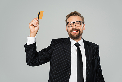 positive businessman in suit and glasses holding credit card isolated on grey