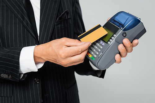 cropped view of businessman in suit holding credit card near payment terminal isolated on grey