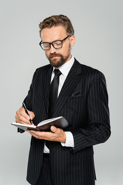 well dressed businessman in glasses and formal wear writing in notebook isolated on grey