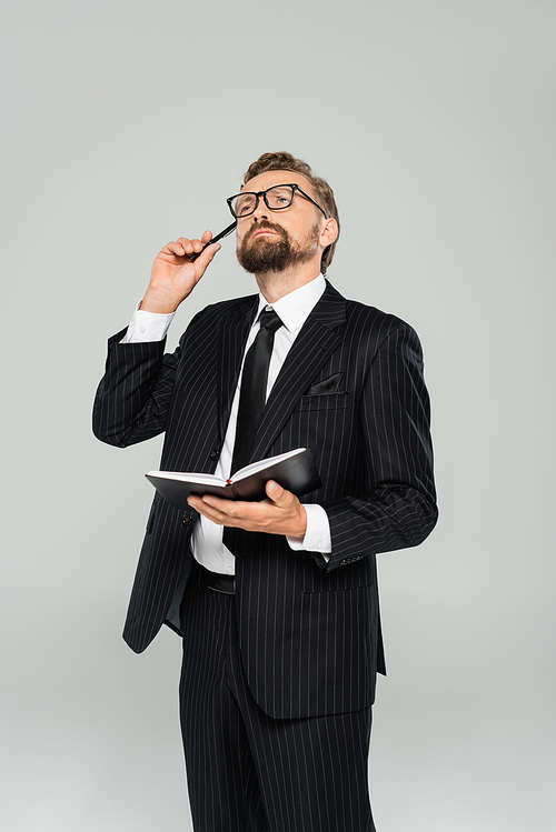 pensive businessman in glasses and formal wear holding notebook isolated on grey