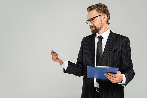 well dressed businessman in glasses holding clipboard and looking at paper isolated on grey