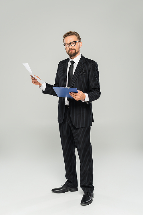 full length of well dressed businessman in glasses holding clipboard and paper on grey