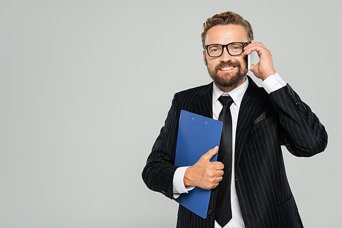 happy businessman in glasses holding clipboard and talking on smartphone isolated on grey