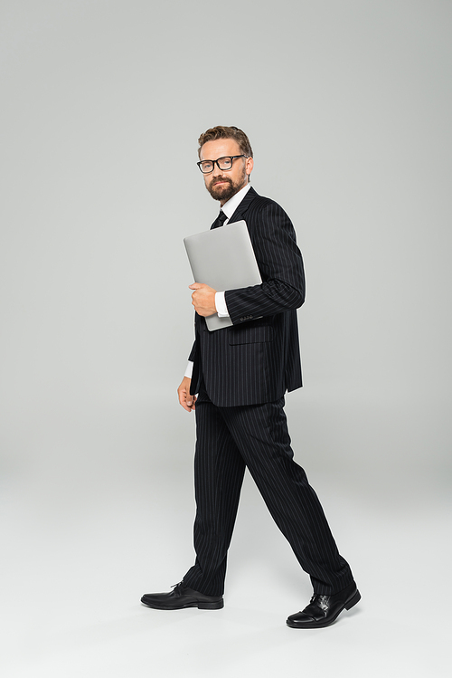 full length of well dressed businessman in glasses walking with laptop on grey