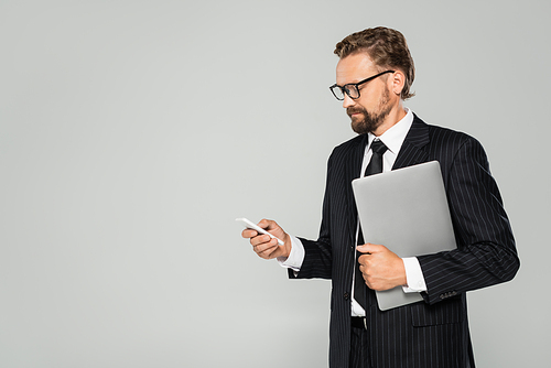 well dressed businessman in glasses holding laptop and using smartphone isolated on grey