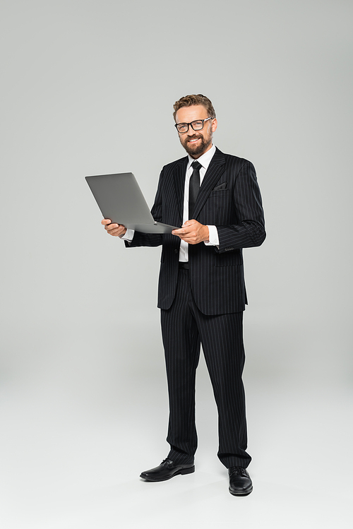 full length of happy and well dressed businessman in glasses using laptop on grey