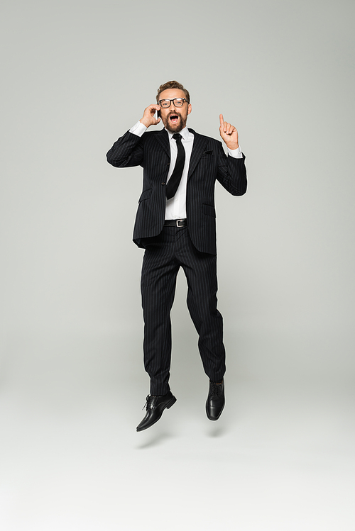 full length of excited businessman in glasses talking on smartphone and pointing up while jumping on grey