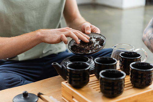 cropped view of man adding puer tea in traditional japanese teapot near cups