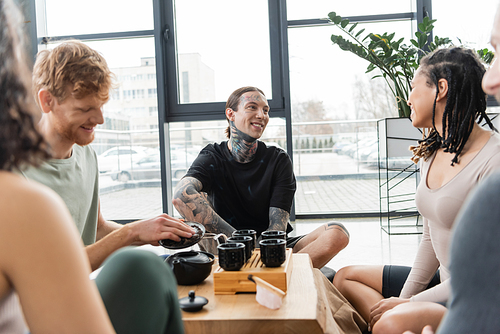 cheerful man with tattoos sitting near interracial friends during tea ceremony in yoga studio