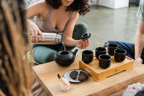 cropped view of woman pouring hot water from thermos while brewing pu-erh tea near people in yoga studio
